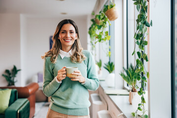 Portrait of a pretty businesswoman, holding a cup of coffee, standing at the home office, smiling.