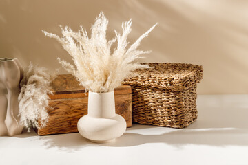 Beige ceramic vase with pampas grass on the table with wooden box organizer and wicker box at the background, cozy sunlight. Scandinavian home decoration, natural eco materials