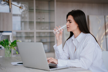Confused young brunette young woman in white shirt sitting at desk with laptop remote works drinks water. Focused student girl tired of distant learns. Exhausted  businesswoman reads news via internet