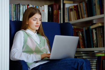 Student sitting in armchair between bookcases and typing on laptop. Young female browsing internet for studying in library. Concept of education