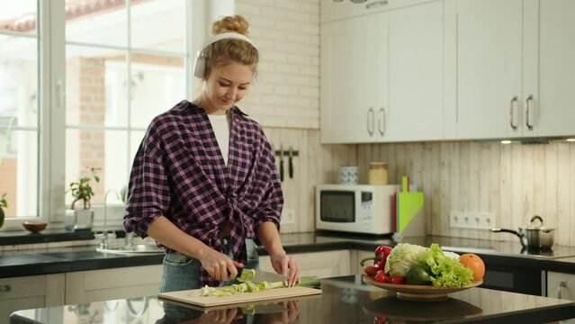 A smiling Caucasian woman wearing headphones is cutting leeks on a wooden cutting board in the kitchen. In front of her on a plate are fresh vegetables. High-quality 4k footage