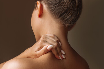 Cropped shot of a young woman standing with her back holds her neck massaging a trapezius muscle with her hand isolated on dark brown background. The girl suffers from an osteochondrosis - obrazy, fototapety, plakaty