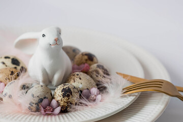 Banner. The concept of a bright holiday is a white rabbit and Easter quail eggs with pink flowers and feathers in a plate on a white isolated background. Beautiful Easter card.