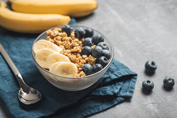 Granola cereal oatmeal with blueberries and banana fruits in a bowl on a blue napkin and grey background