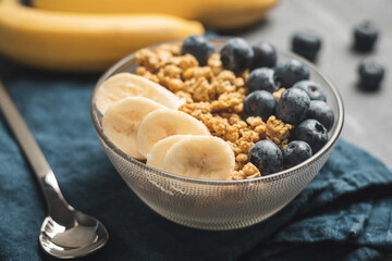 Granola cereal oatmeal with blueberries and banana fruits in a bowl on a blue napkin and grey background