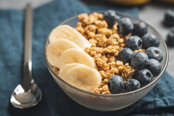 Granola cereal oatmeal with blueberries and banana fruits in a bowl on a blue napkin and grey background