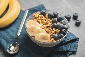 Granola cereal oatmeal with blueberries and banana fruits in a bowl on a blue napkin and grey background