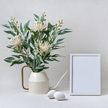 Elegant minimalistic composition with flower bouquet in watering can, eggs and empty white photo frame on light neutral background, spring Easter holiday template with copy space