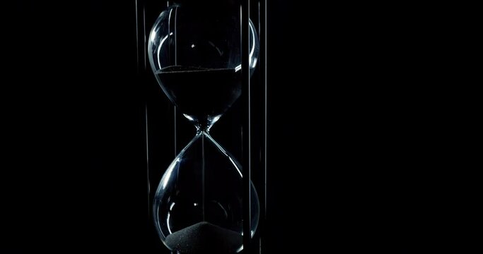 Rotation of sand timer clock with sand flowing from through hourglass on black background., Tilt down
