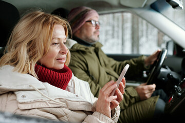 Blond senior woman in winterwear texting in smartphone or scrolling through navigation map while sitting in car against her husband driving