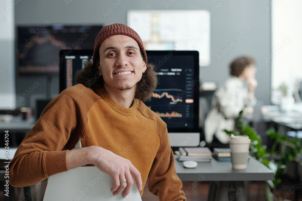 Poster Young smiling IT support manager sitting by workplace in front of camera against computer monitor with graphic data on screen
