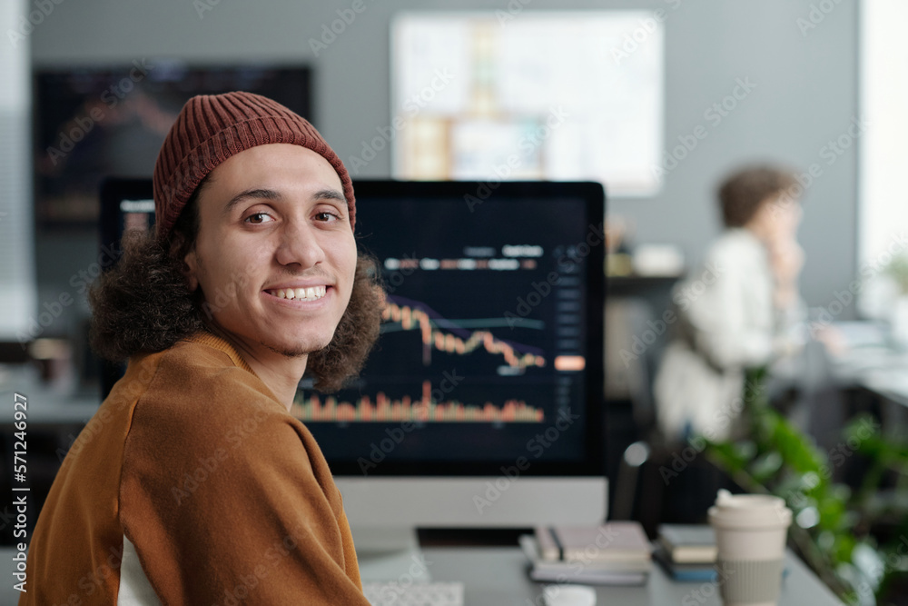 Poster Happy young IT support manager looking at camera while sitting by workplace in front of computer screen with graphic data