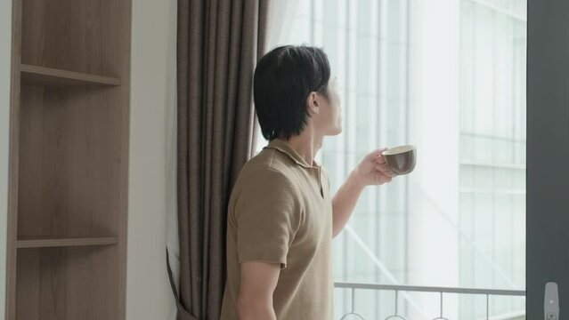 Guy in casualwear walking towards door to balcony, opening it and enjoying new summer morning while standing by wall in bedroom and having cup of tea or coffee