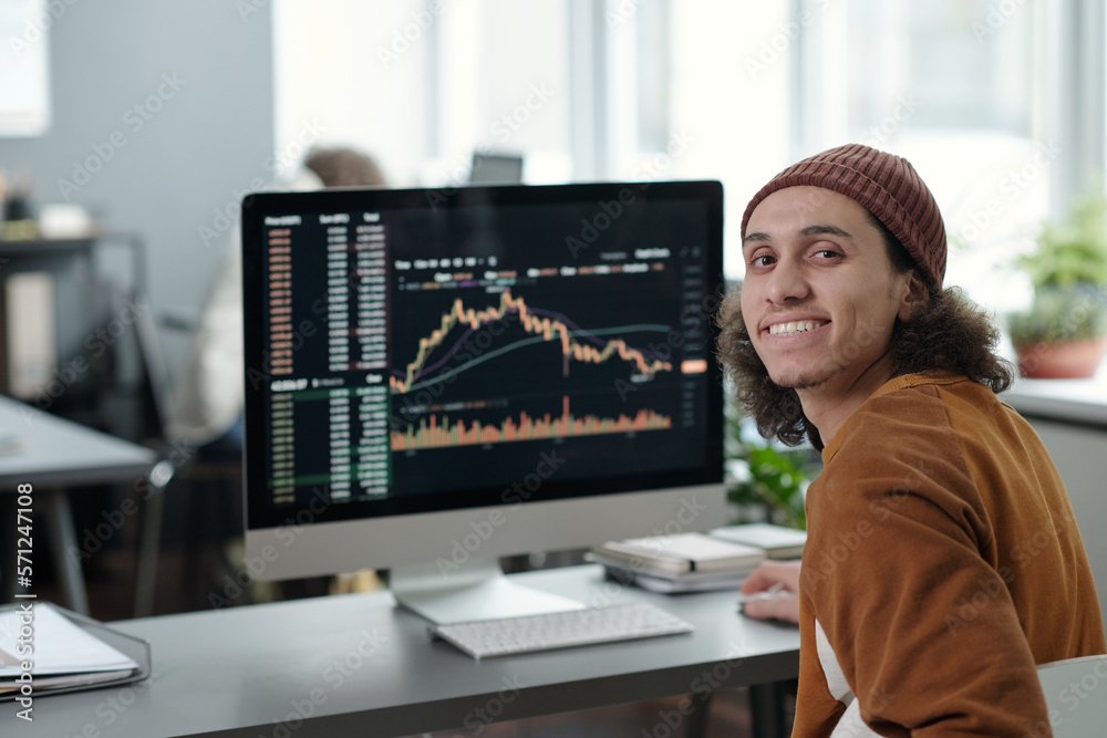 Poster Happy young male IT manager or analyst sitting in front of camera by desk with computer and graphic data on screen and looking at you