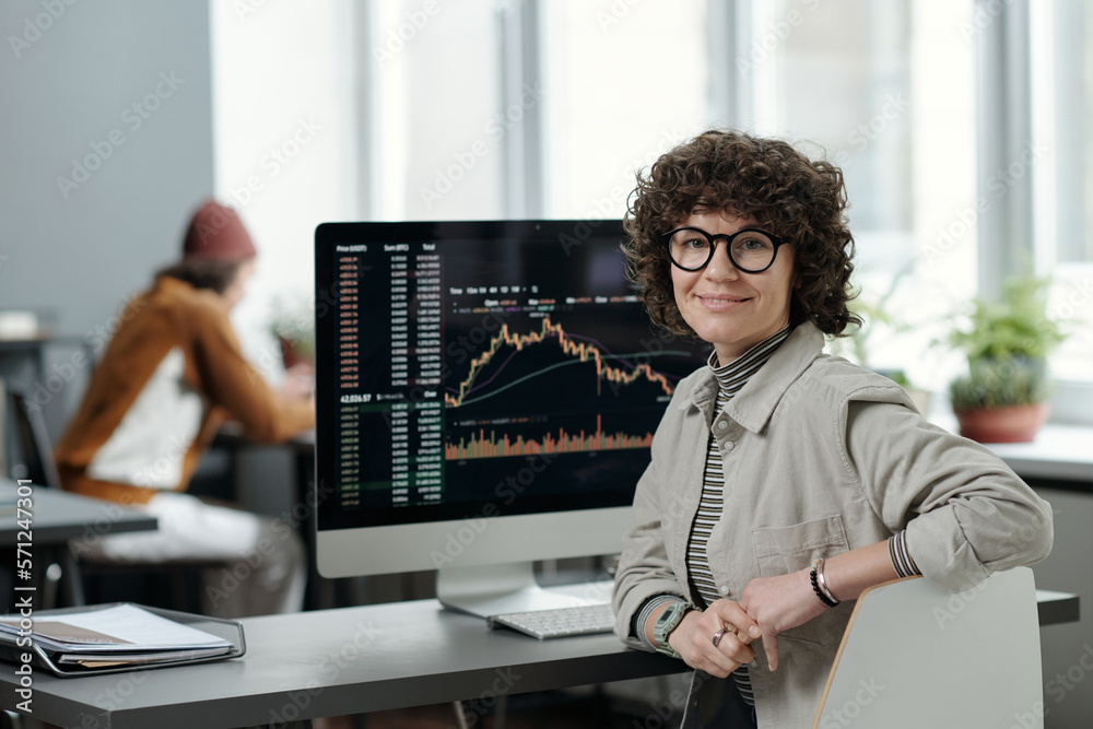 Poster young smiling businesswoman or crypto analyst in casualwear sitting by workplace with computer monit