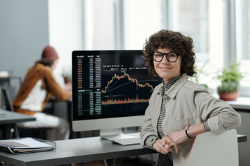 Young smiling businesswoman or crypto analyst in casualwear sitting by workplace with computer...