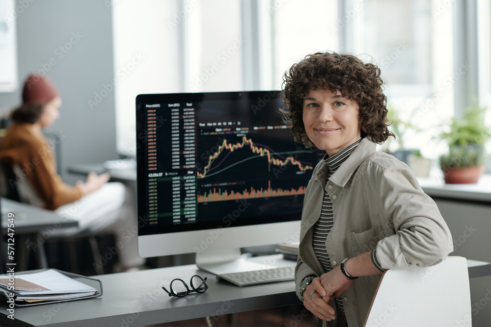 Poster Young smiling female analyst sitting by workplace with computer monitor and looking at you while working with financial graphic data