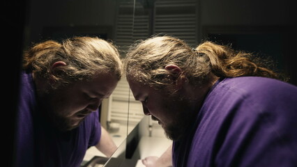 One desperate young overweight man crying in bathroom alone. Desperate tearful person leaning on mirror reflection feeling grief