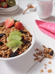 
A bowl of granola. Completed with dried fruit, kiwi, strawberry, honey, honey dipper, mug jug, spoon. Healthy food concept. Isolated background in white.