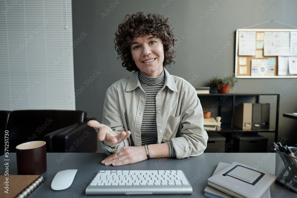 Poster young smiling female manager in casualwear sitting by desk in front of computer moniot, looking at s