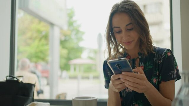 close up beautiful cheerful young woman sits at window in cafe and uses phone for chatting, texting, posting in social media. portrait of attractive caucasian female brunette with smartphone