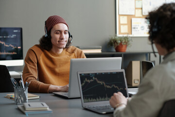 Young smiling male operator in headset and casualwear consulting online clients while sitting by workplace in office in front of laptop