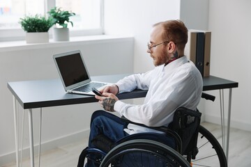 A man wheelchair in the office works at a laptop with a phone, working online, social networks and...