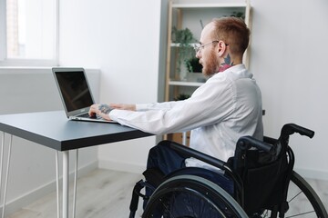 A man in a wheelchair looks at the camera businessman in the office working on a laptop online,...