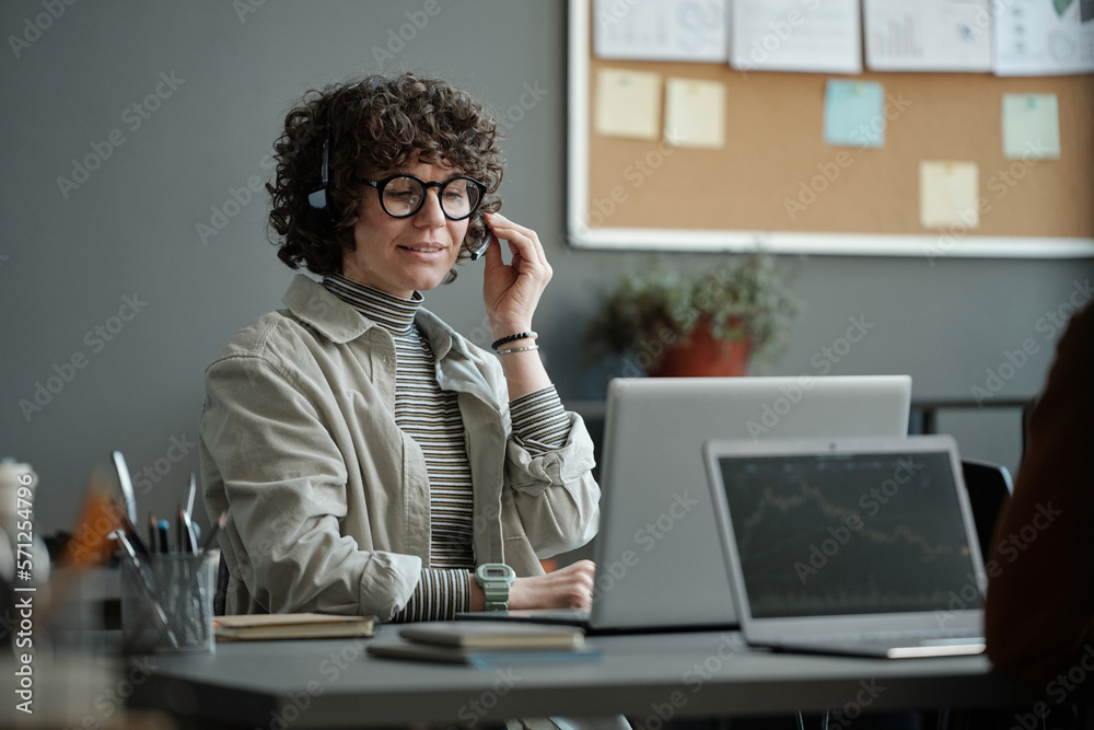 Poster young confident female operator in headset sitting by workplace and talking to online client and hel