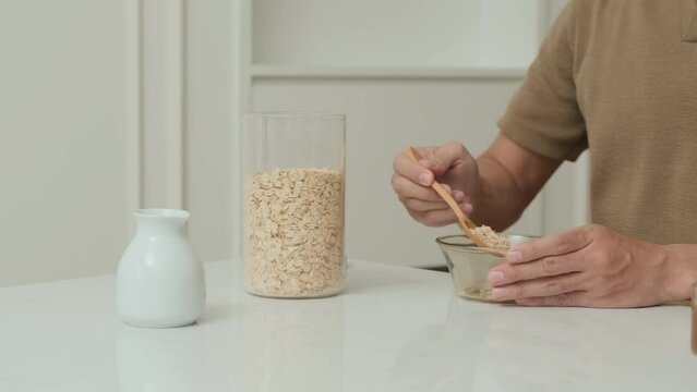Close-up of guy in shirt sitting by kitchen table, taking some oat flakes from glass with wooden spoon and putting it into bowl while preparing healthy and low calorie breakfast