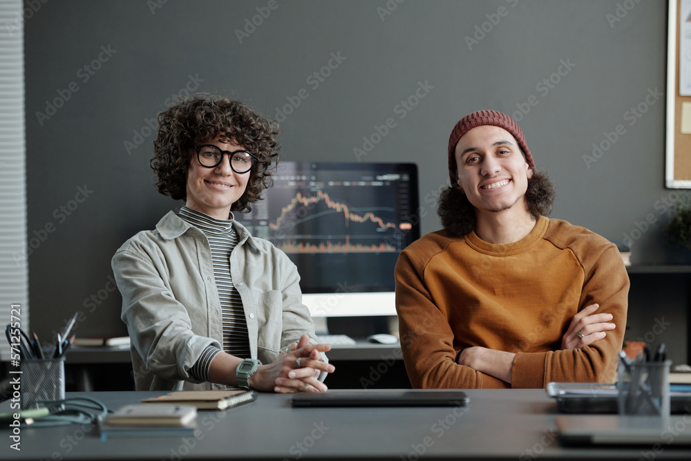 Poster two young intercultural managers in casualwear sitting by workplace in office and looking at camera 