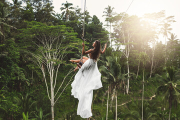 Woman wearing white dress swinging on rope swings with beautiful view on rice terraces and palm...