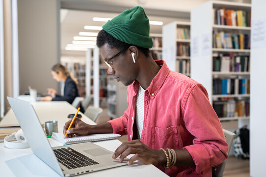 Focused African American Student Online Learner Taking Notes During Webinar, Studying Remotely In Library. Young Black Guy Watching Educational Video Or Lecture On Laptop. Remote Study Concept