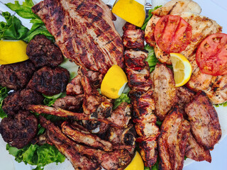 Platter of various types of meat including, pork, lamb, meatballs, and chicken served with lemon and tomato slices close-up top view.