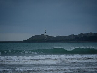 Waves crashing onto ocean beach with black and white Cape Campbell lighthouse in background, Marlborough New Zealand