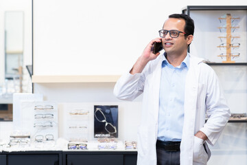 Indian middle aged ophthalmologist or optometrist man work at optometry clinic, using mobile phone appointing patient to check eyesight and vision or optical consulting