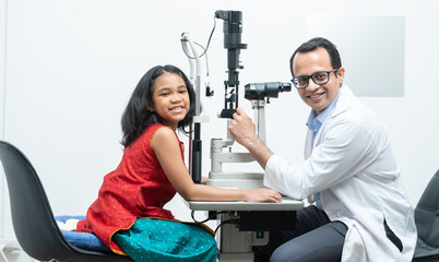 Indian middle aged professional ophthalmologist or optometrist man work at optometry clinic, using optometry instruments little girl patient to check eyesight and eyes vision. smile at camera