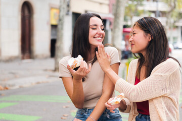 two young friends laughing and having fun while sharing some sweet buns sitting in a city park,...