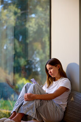 Depressed Teenage Girl Sitting By Window At Home Looking At Mobile Phone