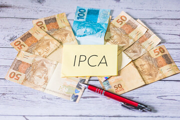 A sigla IPCA de Índice Nacional de Preços ao Consumidor Amplo em Português do Brasil escrita em um pedaço de papel. Notas do Real Brasileiro na composição. Economia brasileira.