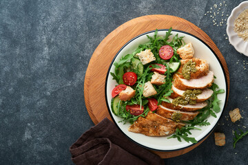 Fresh salad with grilled chicken fillet, lettuce, arugula, spinach, tomatoes, cucumber and pesto sauce, sesame seeds with olive oil in white bowl on black slate background. Healthy lunch menu Top view