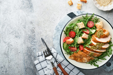 Fresh salad with grilled chicken fillet, arugula, tomatoes, cucumber and pesto sauce, sesame seeds with olive oil in white bowl on light slate background. Healthy lunch menu. Diet food. Top view.