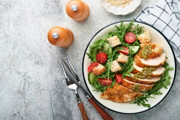 Fresh salad with grilled chicken fillet, arugula, tomatoes, cucumber and pesto sauce, sesame seeds with olive oil in white bowl on light slate background. Healthy lunch menu. Diet food. Top view.
