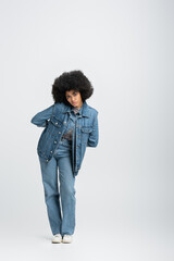 full length of curly african american woman in denim outfit looking at camera while standing on grey.