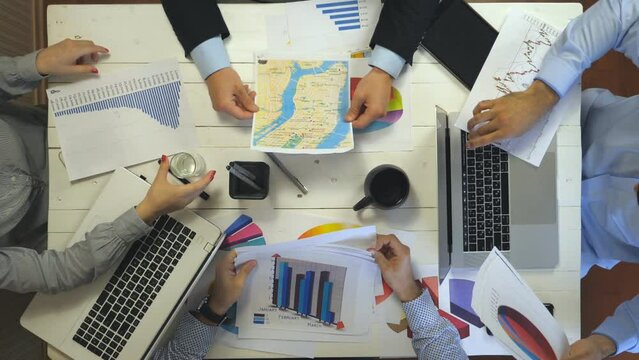 Top view to hands of business people sitting at table in trendy office and examining graphs. Group of busy employees working efficiently on business development. Creative team work indoor. Slow mo