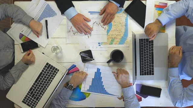 Top view to hands of four business people sitting at table in office and examining graphs. Group of busy employees working efficiently on business development. Creative team work indoor. Slow motion