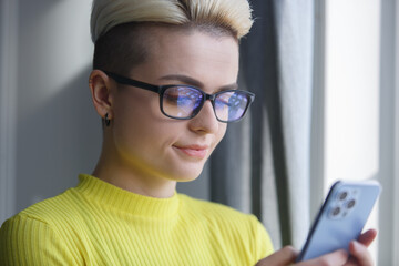 Beautiful white woman with short dyed hair browsing mobile app on modern smart phone. Portrait of stylish tom boy female using mobile phone - obrazy, fototapety, plakaty