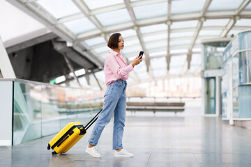 Fototapeta na wymiar Beautiful Young Woman With Suitcase Walking At Airport Terminal And Using Smartphone