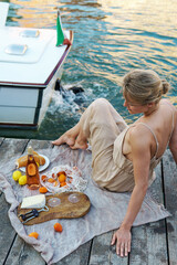 Lovely young woman picturesque picnic on the wooden gondola dock with rose wine, fruits and snack on wooden pier