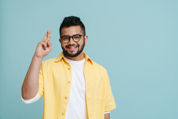 Fototapeta premium Smiling indian man holding fingers crossed while standing isolated over blue wall